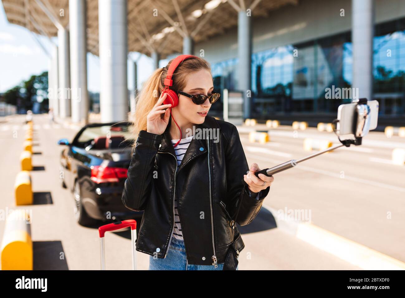 Hübsche Mädchen in Sonnenbrille und Kopfhörer, die Fotos auf dem Handy in der Nähe des Flughafens mit Cabriolet auf dem Hintergrund machen Stockfoto