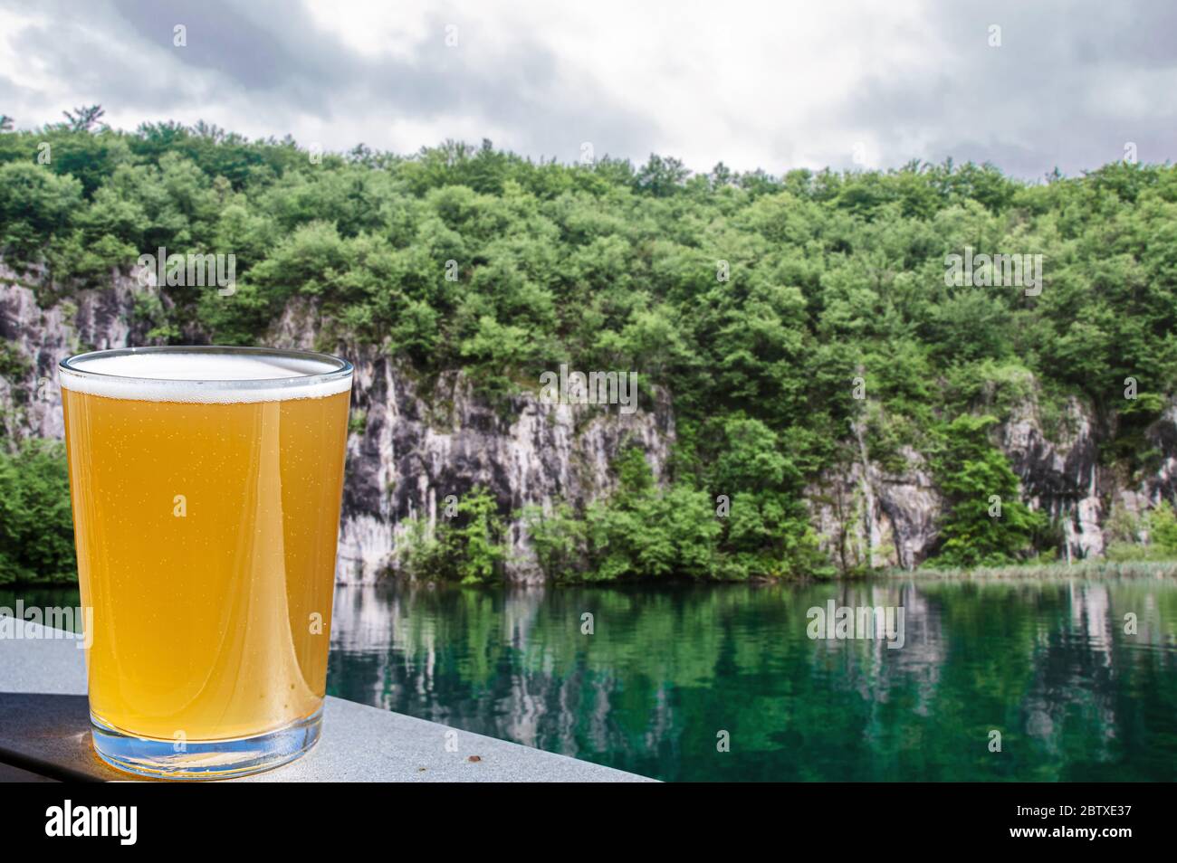 Ein Glas helles Bier mit Blick auf den smaragdgrünen See in Kroatien. Lagerbier gegen steile Ufer mit Bäumen. Stockfoto