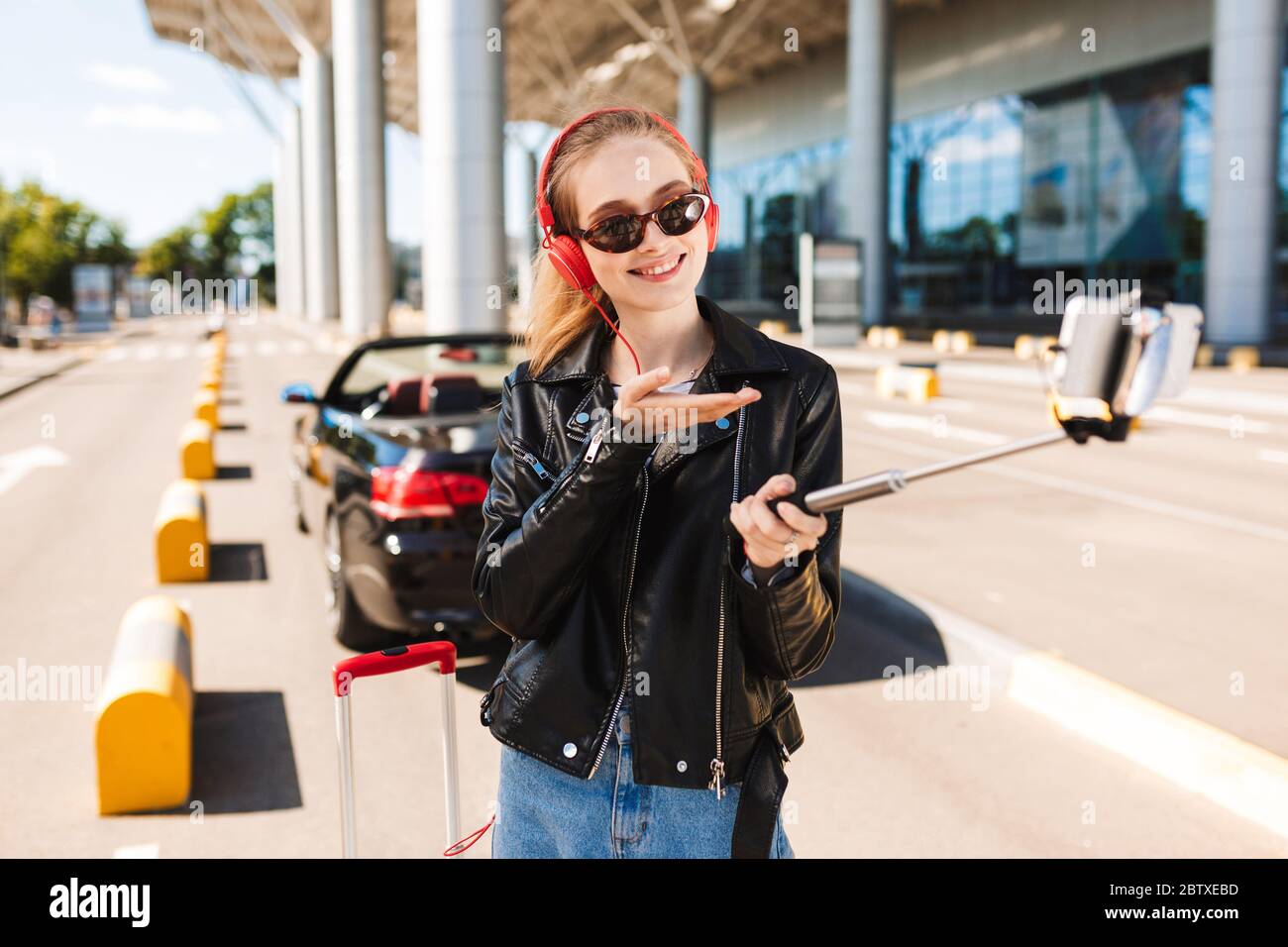 Fröhliches Mädchen in Sonnenbrille und Kopfhörer glücklich Foto auf Handy mit schwarzem Cabriolet auf Hintergrund Stockfoto