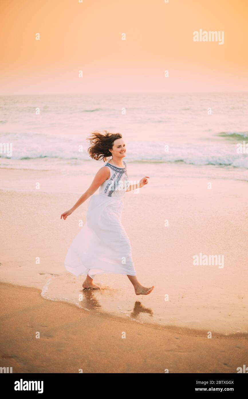Goa, Indien. Junge Kaukasische Frau Mit Flatterndem Haar Im Wind In Weißem Kleid Wandern Entlang Der Seashore, Das Leben Genießen Und Lächeln Im Sommer Sonnenlicht. Stockfoto