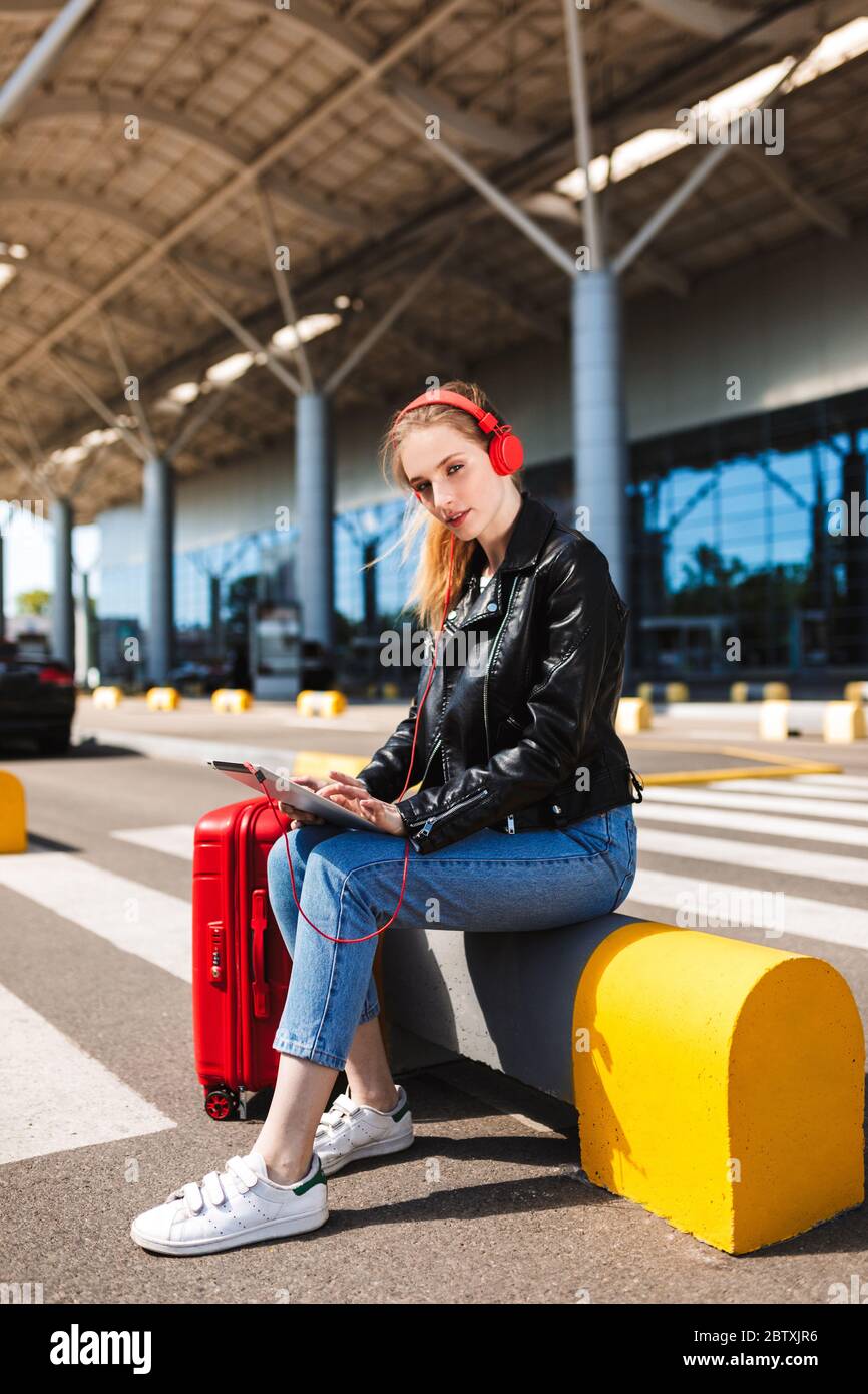 Schönes Mädchen in Kopfhörer traumhaft in der Kamera suchen hält das Tablet in den Händen und Koffer in der Nähe mit Flughafen auf Hintergrund Stockfoto