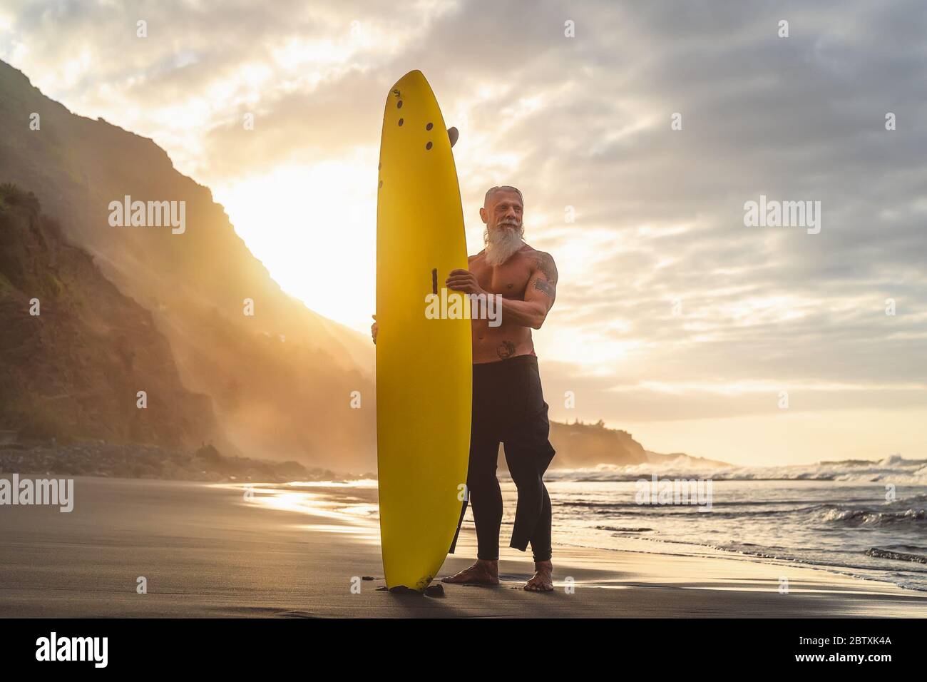 Happy Fit Senior mit Spaß beim Surfen bei Sonnenuntergang - sportliches bärtiger Mann Training mit Surfbrett am Strand Stockfoto