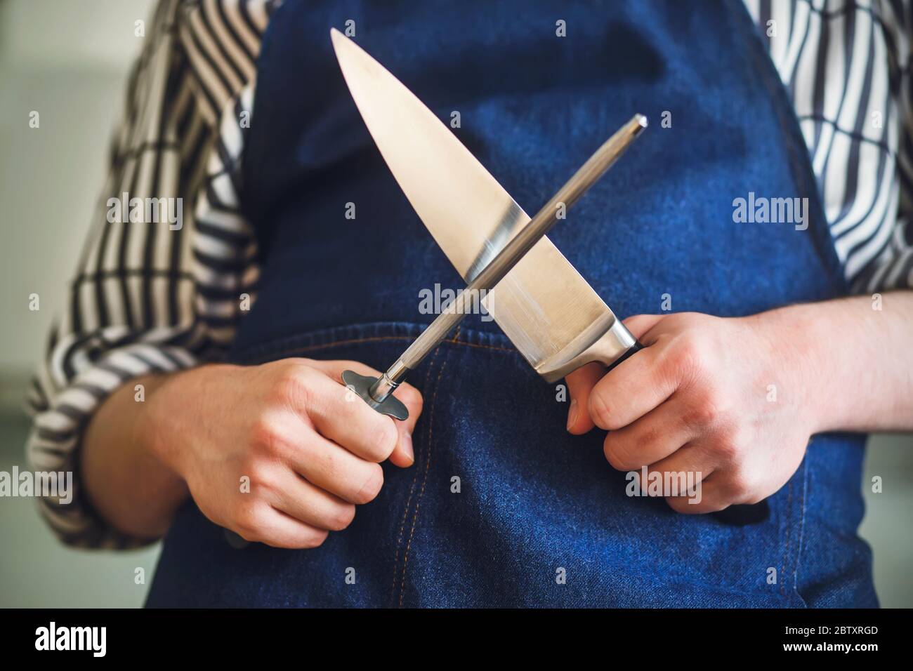 Ein Koch in blauer Schürze und gestreiftem Hemd wird vor dem Kochen ein Küchenmesser auf einem langen Wetzstein schärfen. Stockfoto