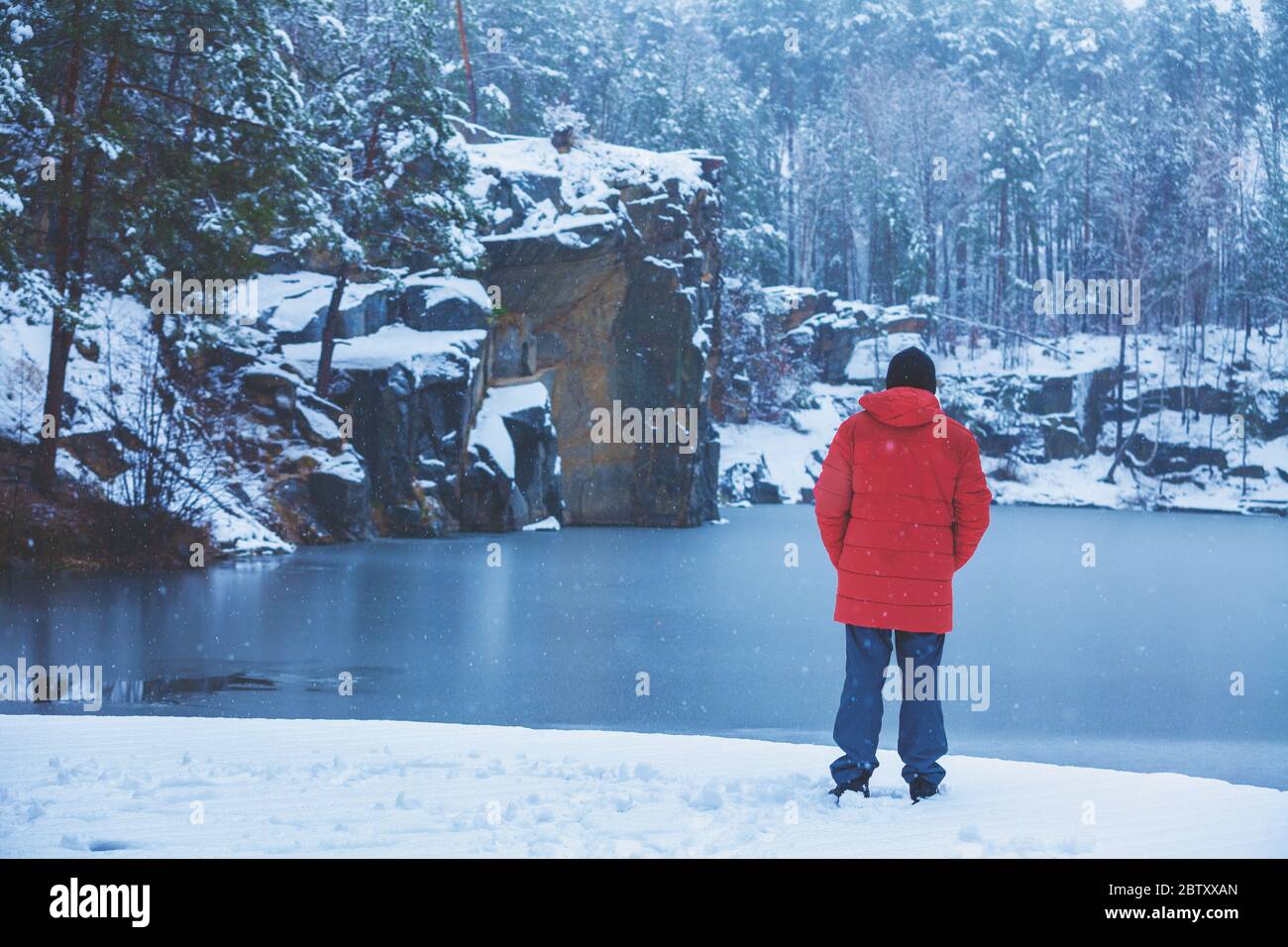 Ein Mann bleibt an einem Seeufer, im Winter mit Schnee bedeckt und schaut auf den schönen See zurück zur Kamera Stockfoto