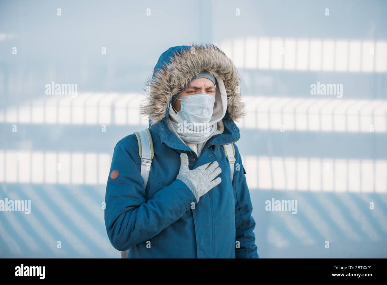 Porträt des kranken jungen Mannes in blauer Jacke auf eine Kapuze, mit einer Erkältung, Gefühl unwohl, trägt medizinische Gesichtsmaske, erleben Brustschmerzen, im Freien. Stockfoto
