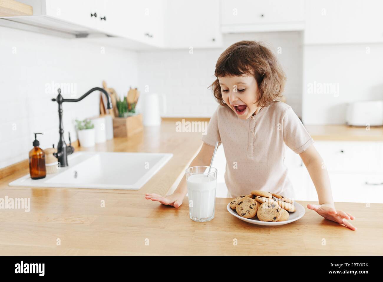 Nettes Kleinkind Mädchen trinken Milch mit Stahlstroh aus Glas und essen Plätzchen sitzen am Küchentisch. Reduzieren Sie die Verwendung von Kunststoff zu Hause mit Kindern Stockfoto