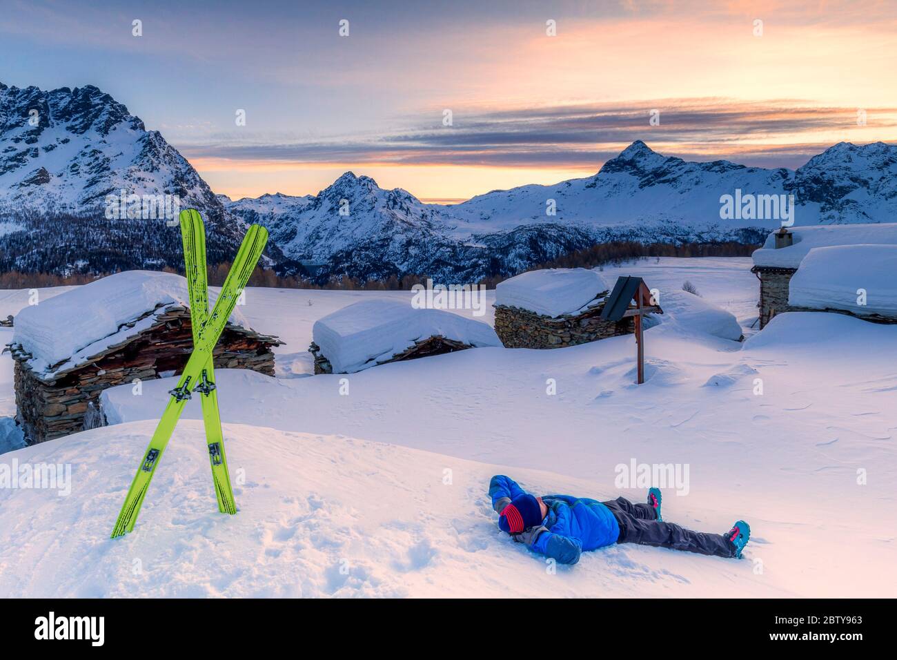 Junge Skifahrer entspannt sich im Schnee mit Blick auf das kleine Dorf bei Sonnenaufgang, Valmalenco, Valtellina, Lombardei, Italien, Europa Stockfoto