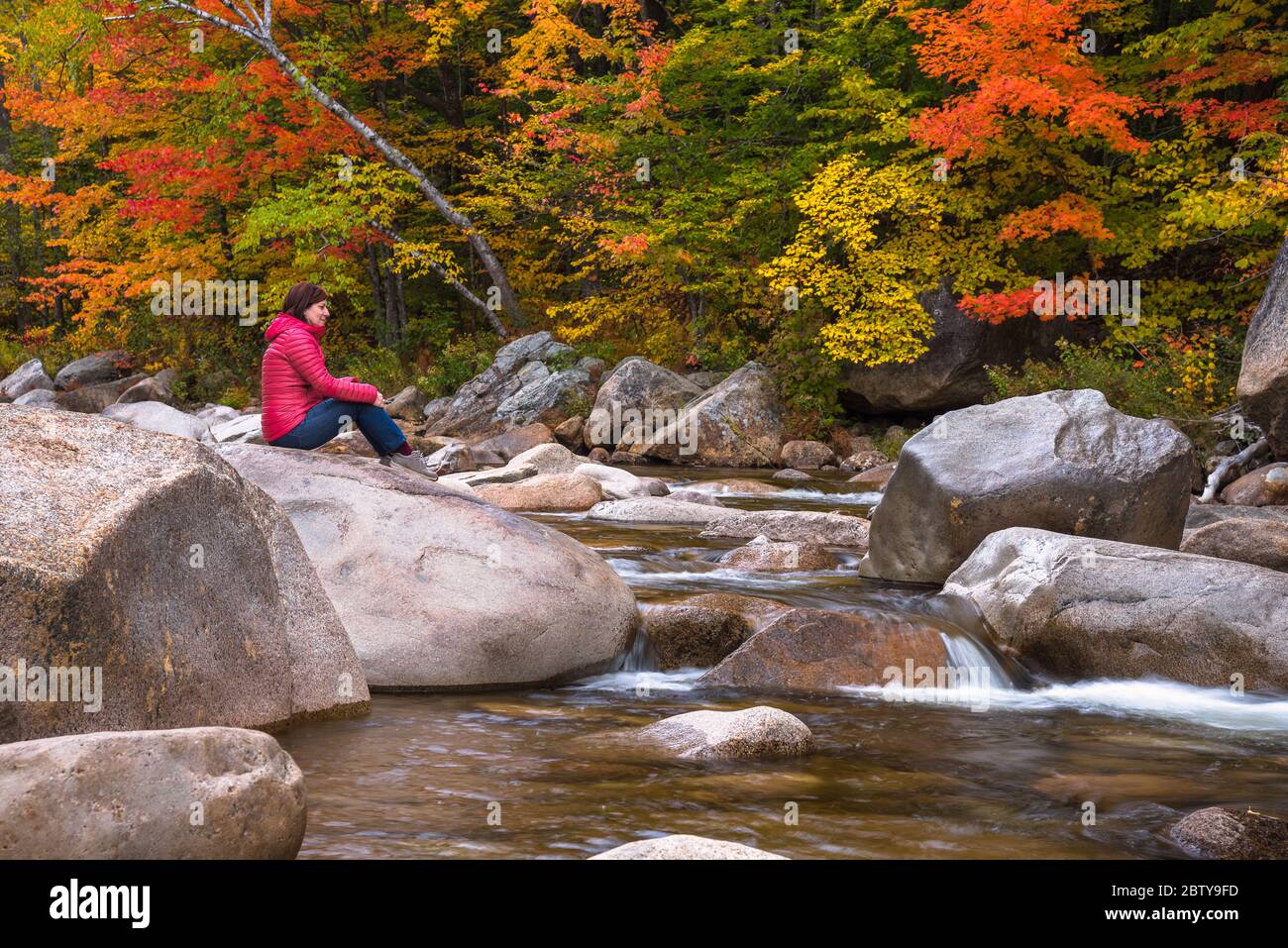 Einsame nachdenkliche Frau sitzt auf einem Felsen auf einem Berg Fluss an einem Herbsttag. Schöne Herbstfarben im Hintergrund. Stockfoto