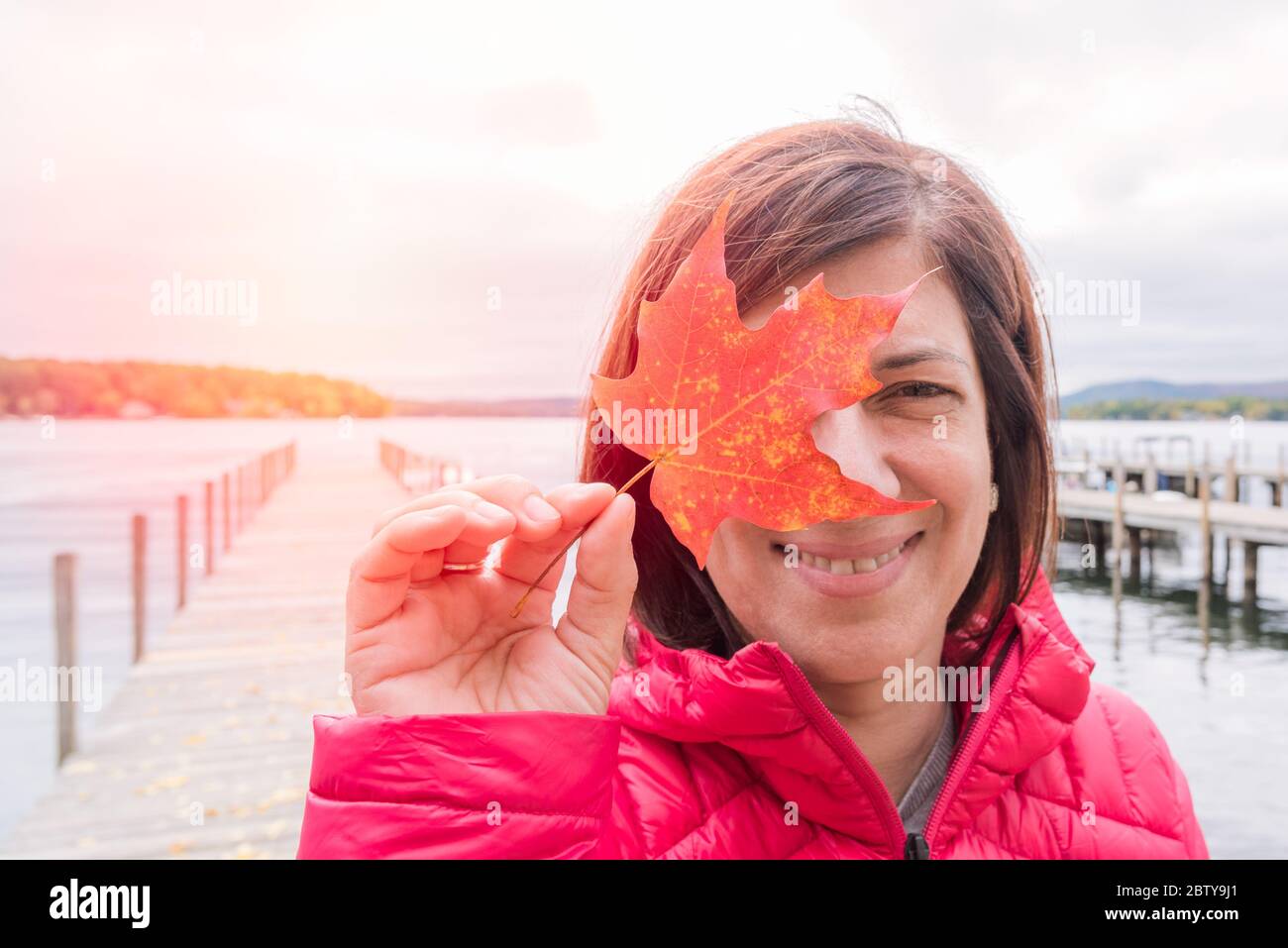 Portrain einer lächelnden Frau, die ein rotes Ahornblatt vor ihrem rechten Auge hält. Im Hintergrund ist ein verlassener Steg auf einem See zu sehen. Stockfoto