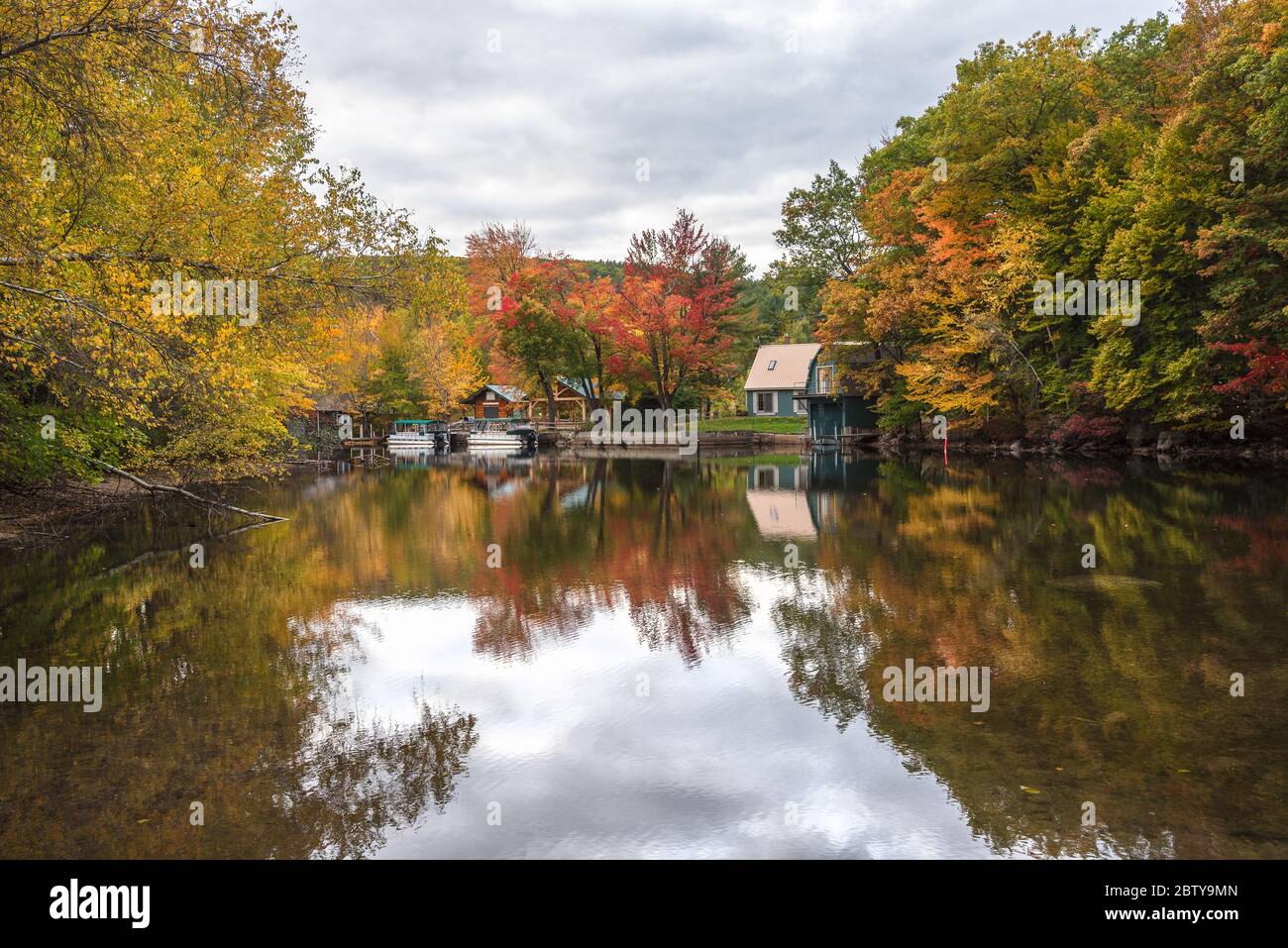 Bootshäuser und Boote entlang eines Flusses mit bewaldeten Ufern an einem bewölkten Herbsttag. Atemberaubende Herbstfarben und Reflektionen im Wasser. Stockfoto