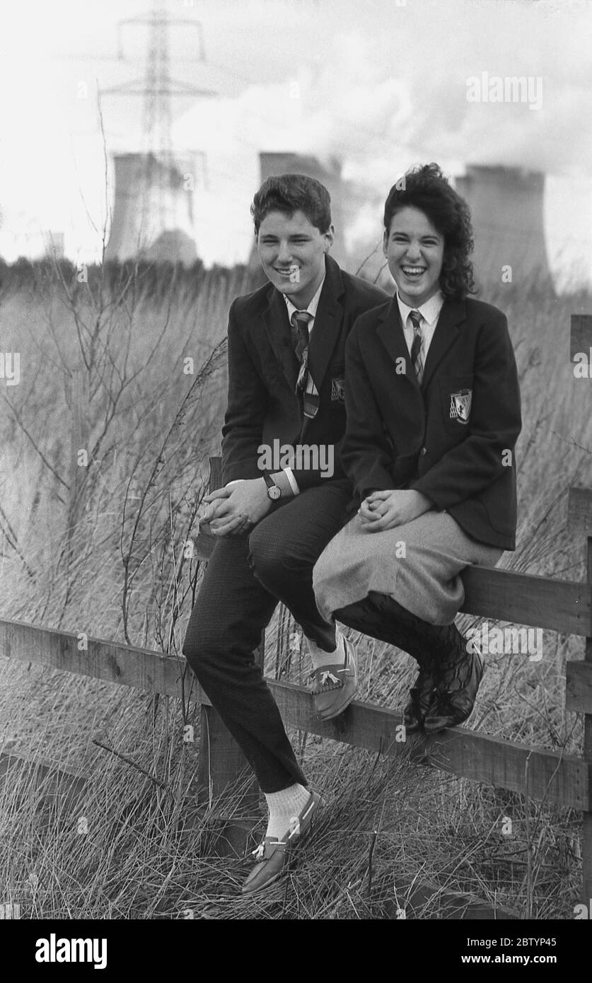 1980, historisch, sitzend auf einem gebrochenen Zaun und lachend, zwei junge Erwachsene, Sechstbildner in Schuluniform, die sich auf einem "Auszeit" von der Schule amüsieren, nachdem sie ein lokales Kraftwerk als Teil ihrer Kursarbeit besucht haben, England, Großbritannien. Stockfoto
