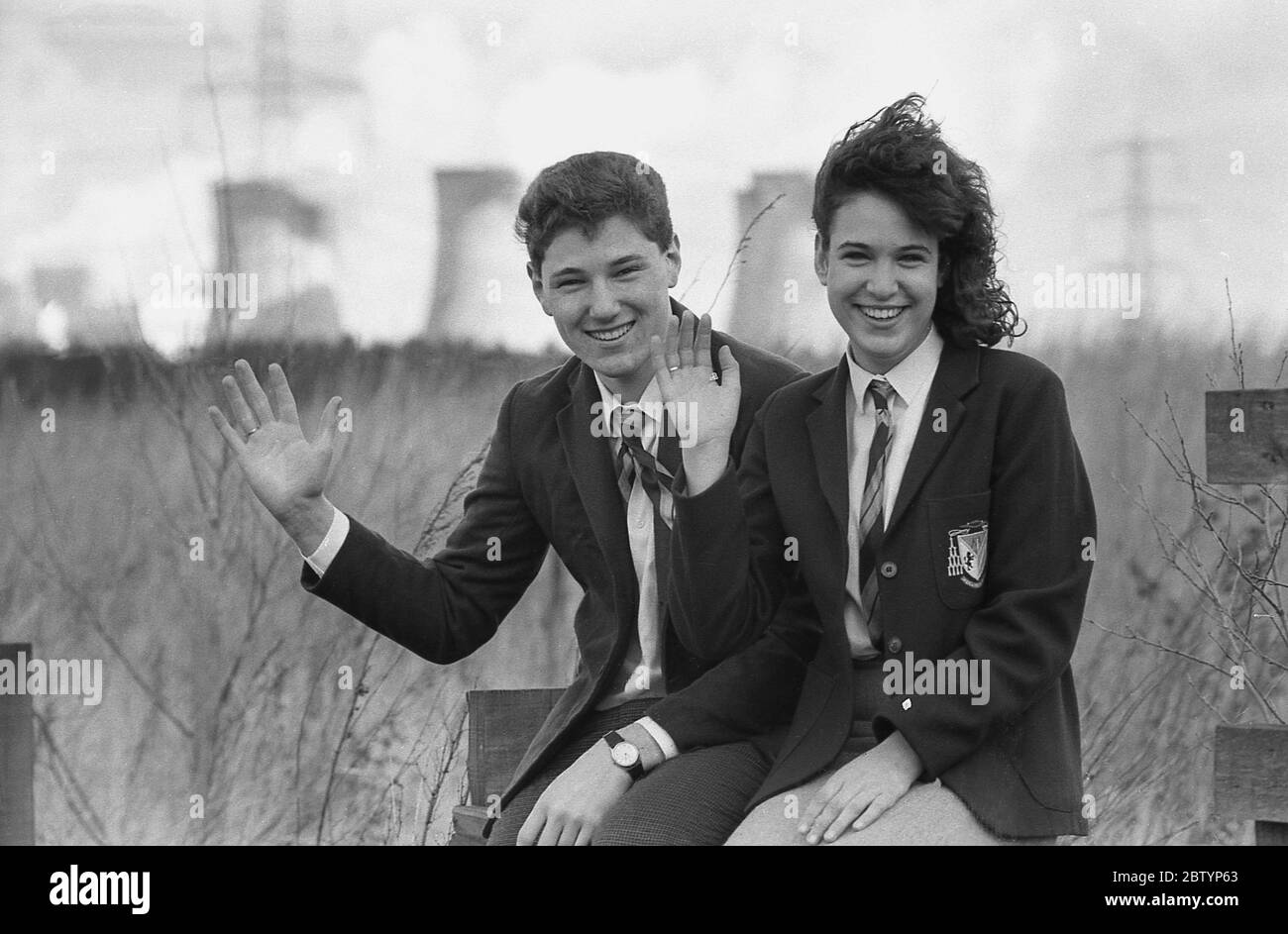 1980, historisch, auf einem Holzzaun sitzend, mit großem Lächeln und winkenden, zwei Sechstbildner in Schuluniform, die sich auf einem "Tag-out" von der Schule amüsieren, nachdem sie im Rahmen ihres Studiums ein lokales Kraftwerk besucht haben, England, Großbritannien. Stockfoto