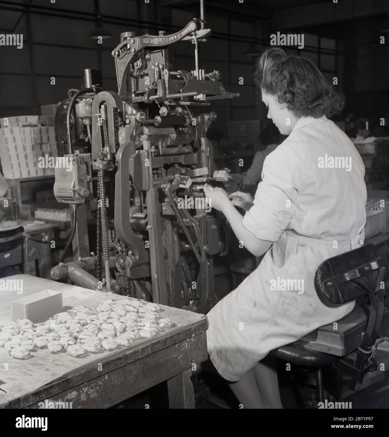 1950er Jahre, historisch, eine Fabrikarbeiterin in einem weißen Mantel sitzt auf einem Metallhocker mit einer industriellen Etikettiermaschine für Flaschen von Pillen, bei der medizinischen Herstellung und Versorgung Unternehmen, Arthur H. Cox's aus Brighton, England, Großbritannien. Stockfoto