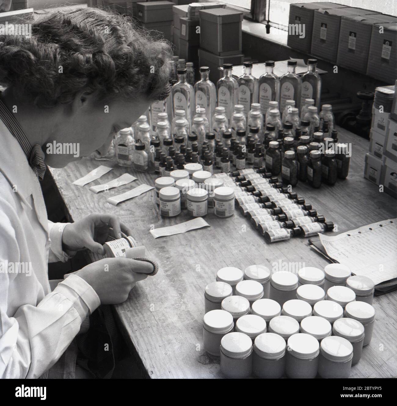 1950er Jahre, historisch, eine Arbeiterin in einem weißen Mantel sitzt an einer Werkbank Etikettierung Flaschen mit medizinischen Pillen oder Tabletten, bei der pharmazeutischen Herstellung und liefert Firma, Arthur H. Cox's aus Brighton, England, Großbritannien. Der Firmengründer Arthur H. Cox erfand die Beschichtung von Tabletten. Stockfoto