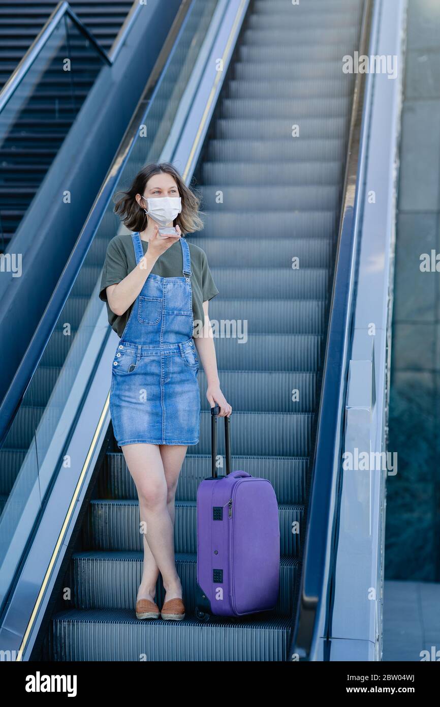 Frau mit Schutzmaske an der Rolltreppe, die ihren Koffer und ihr Telefon hält Stockfoto