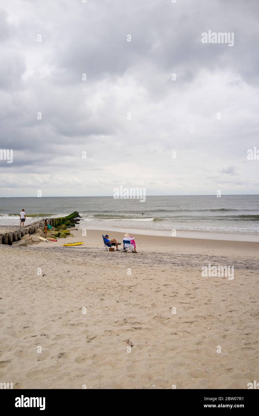 Altes Paar, das in Strandliegen an der Küste von Jersey sitzt. Mit Mann, der auf dem Steg läuft. Wolkiger Tag, viel Sand und Himmel für Kopierraum Stockfoto
