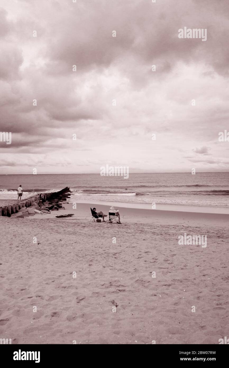 Altes Paar, das in Strandliegen an der Küste von Jersey sitzt. Mit Mann, der auf dem Steg läuft. Wolkiger Tag, viel Sand und Himmel für Kopierraum Stockfoto