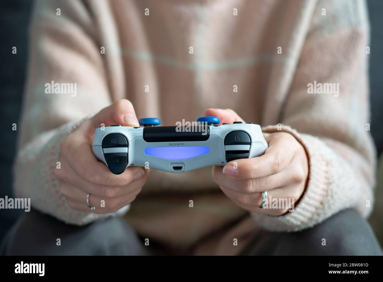 Frau, die in legerer, bequemer Kleidung sitzt, auf dem Sofa sitzt und Videospiele mit dem weißen Sony Wireless-Controller spielt. Stockfoto
