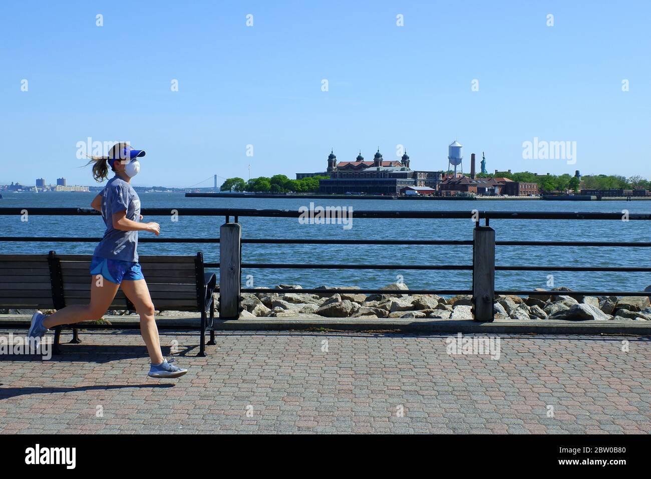 Eine weibliche Joggerin trägt Maske Joggen auf Hudson River Waterfront Walkway mit Ellis Island im Hintergrund.Liberty State Park.Jersey City.New Jersey.USA Stockfoto