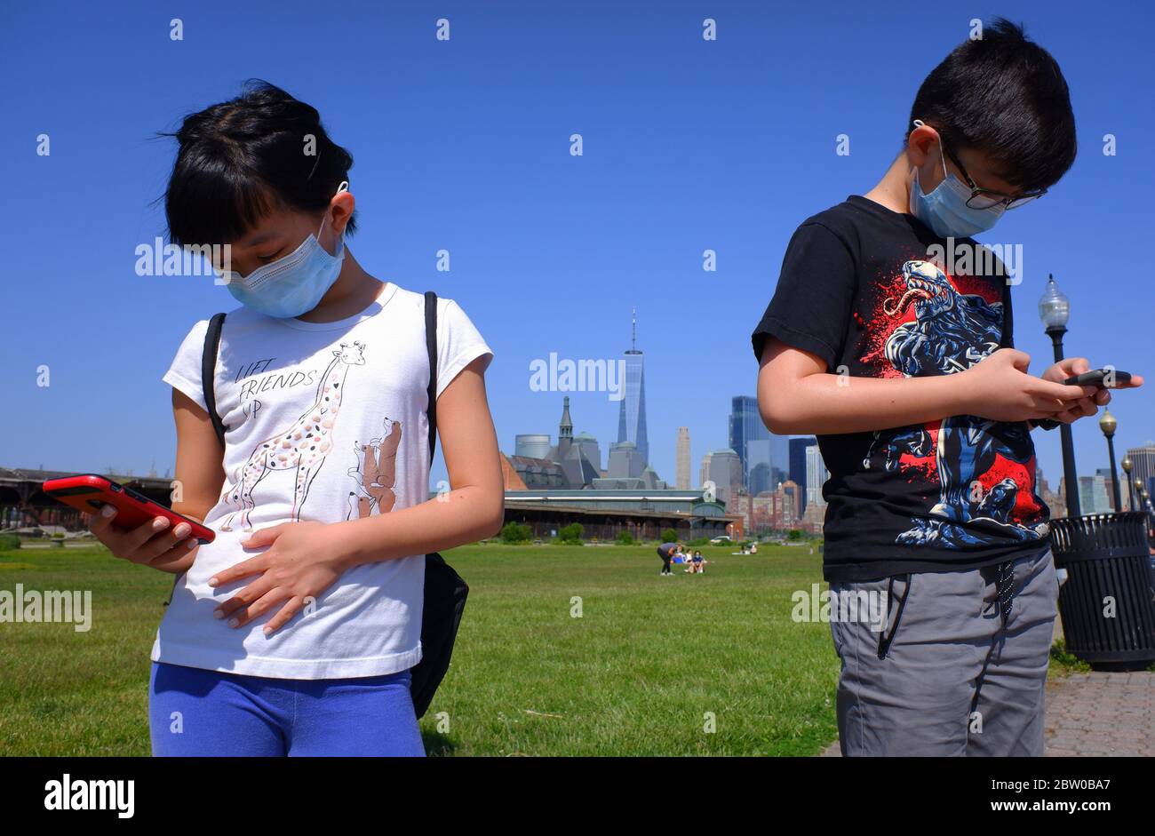 Junge asiatische Mädchen und Jungen tragen Maske Spiele auf dem Handy im Liberty State Park während des Covid-19 Coronavirus Ausbruch im Liberty State Park mit der Skyline von Lower Manhattan New York City Financial District im Hintergrund. New Jersey.USA Stockfoto