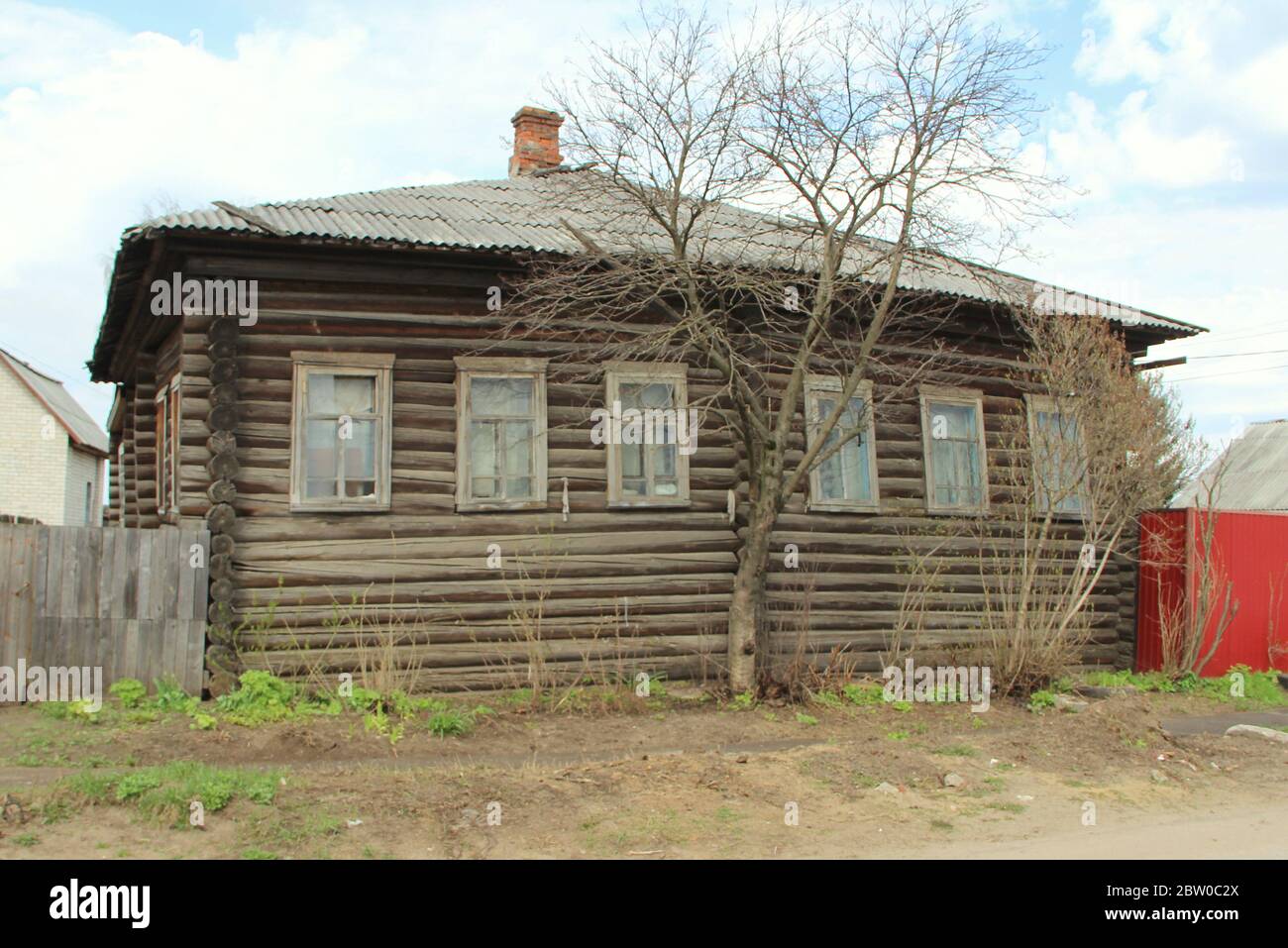 Ein altes Dorfholzhaus mit Schieferdach und einem Ziegelkamin. Der Baum wächst vor dem Haus. Stockfoto