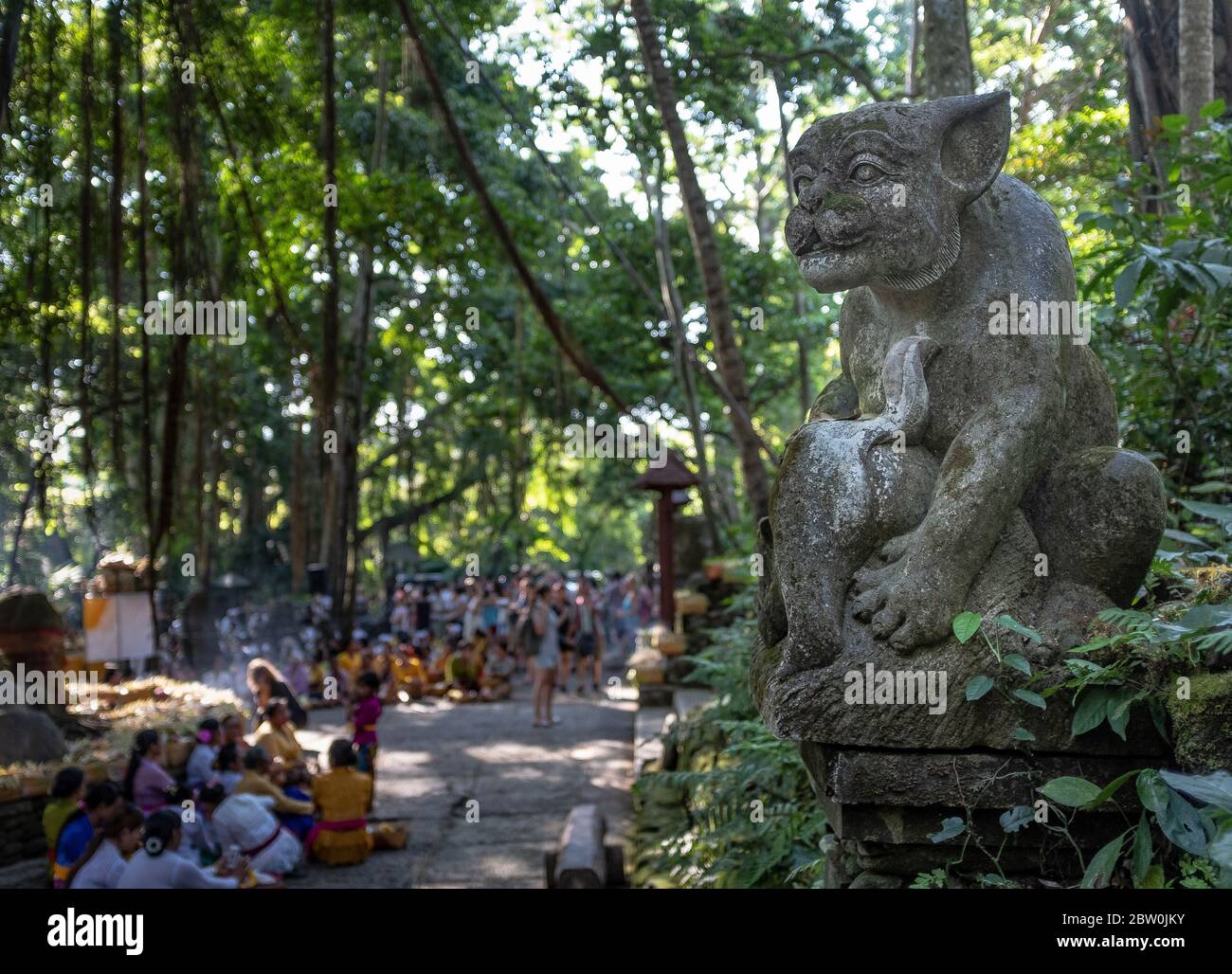 Skulptur einer Katze und eine verschwommene fortlaufende traditionelle Zeremonie im Hintergrund, im Monkey Forest Park, Ubud, Bali, Indonesien Stockfoto