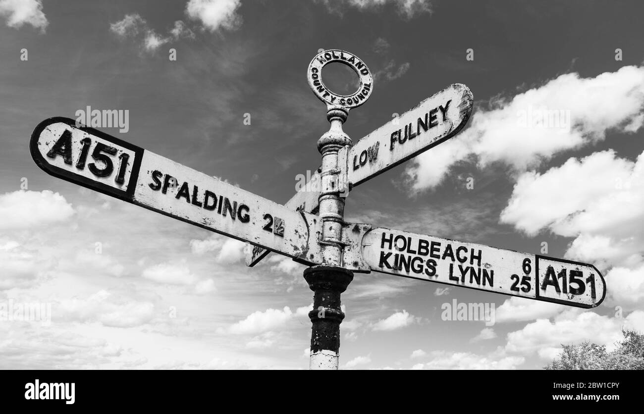 Ein altmodisches britisches Straßenschild auf der A151, das auf Spalding, Holbeach, Kings Lynn und Low Fulney Estate in schwarz und weiß vor Wolken zeigt Stockfoto