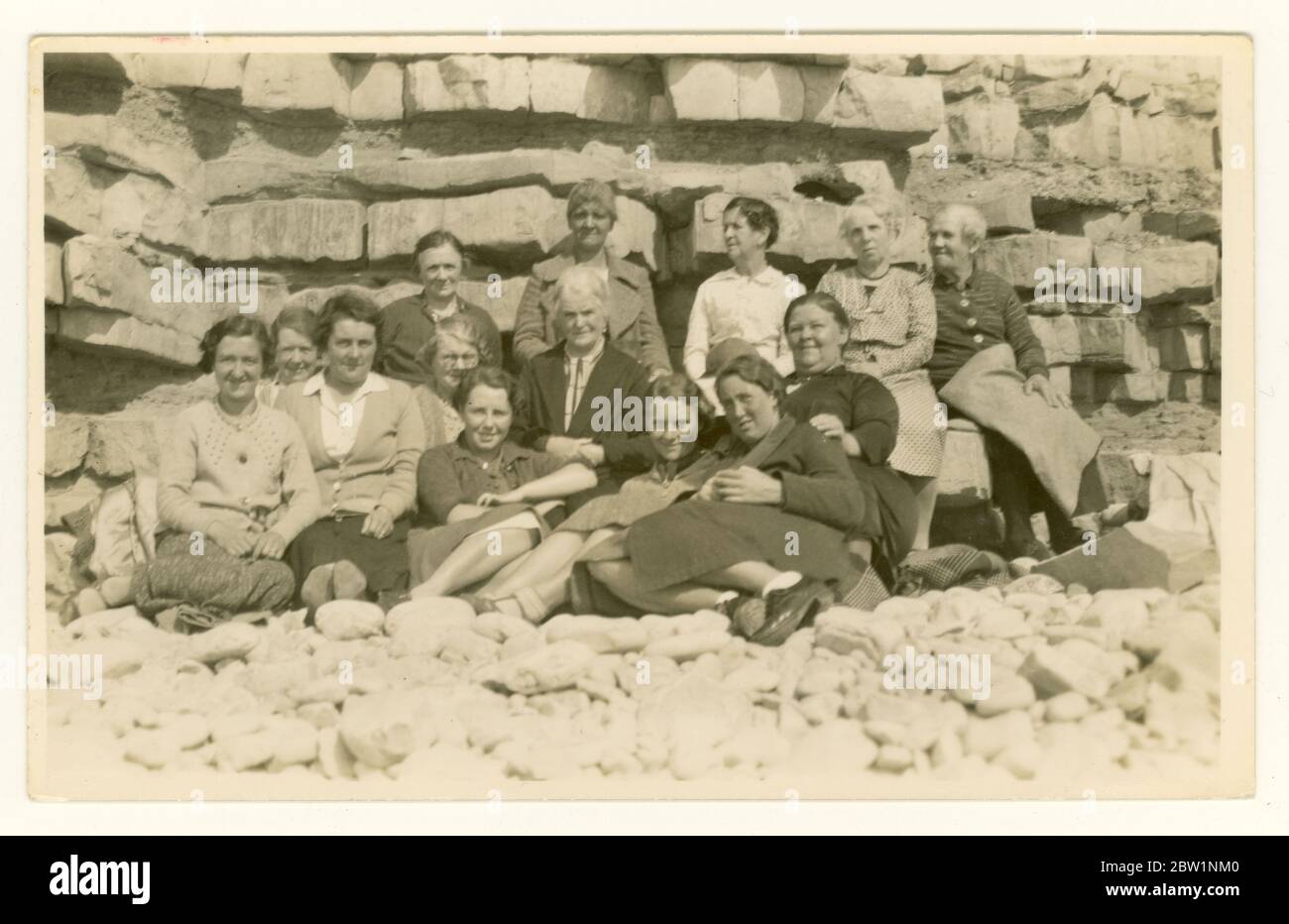 Anfang der 1900er Jahre Postkarte mit einer großen Gruppe älterer Menschen, altmodische Kleidung, freche Tagesausflügler, die am Strand im Schutz einer felsigen Klippe sitzen, um die 1930er Jahre, UK Retro Beach Foto. Stockfoto