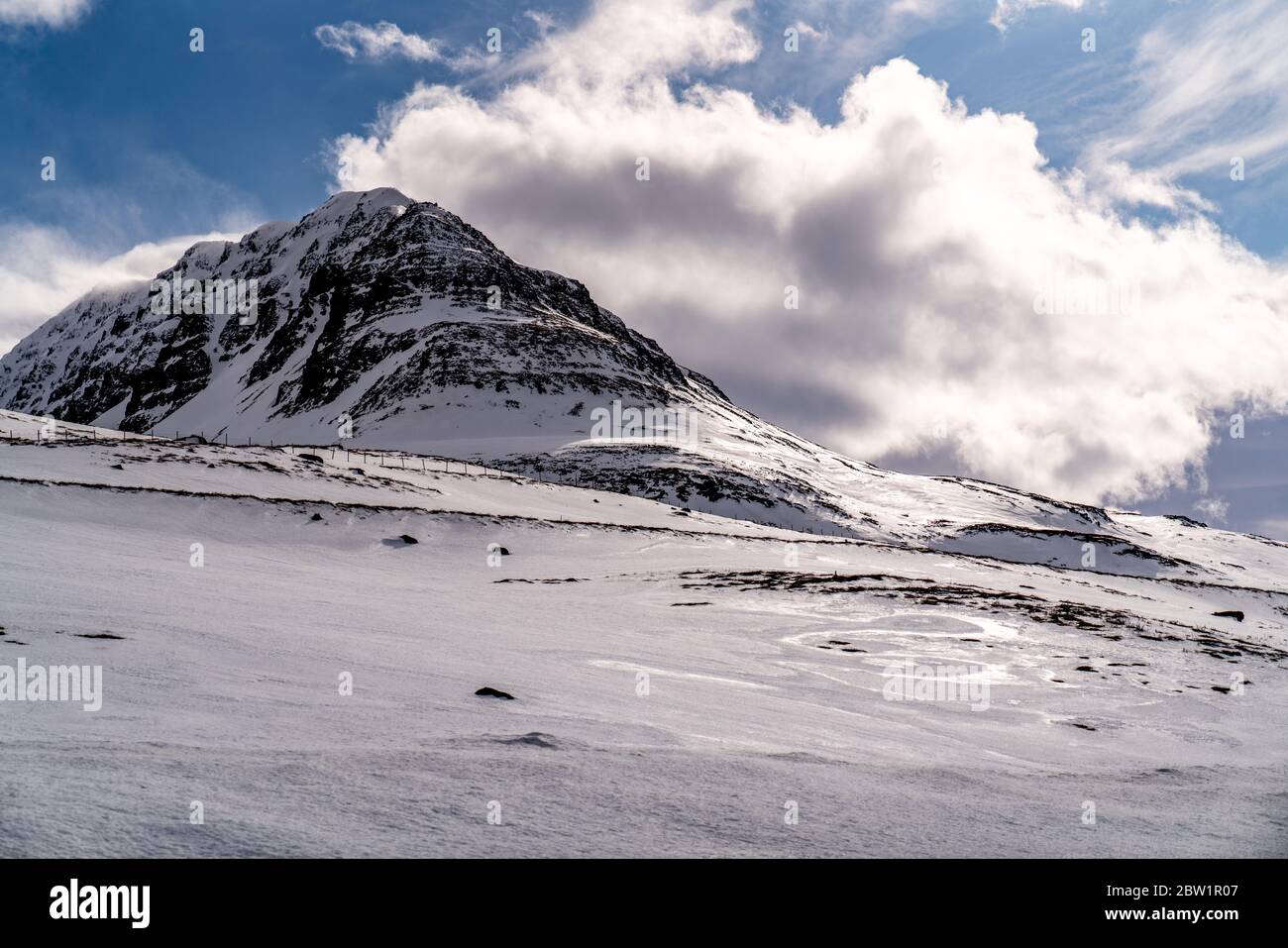 Ein Berggipfel, der an einem sonnigen Tag in Island nach den wirbelnden Wolken gegen einen blauen Himmel greift. Die Sonne reflektiert sich in den eiskigen Teilen des Schnees. Stockfoto