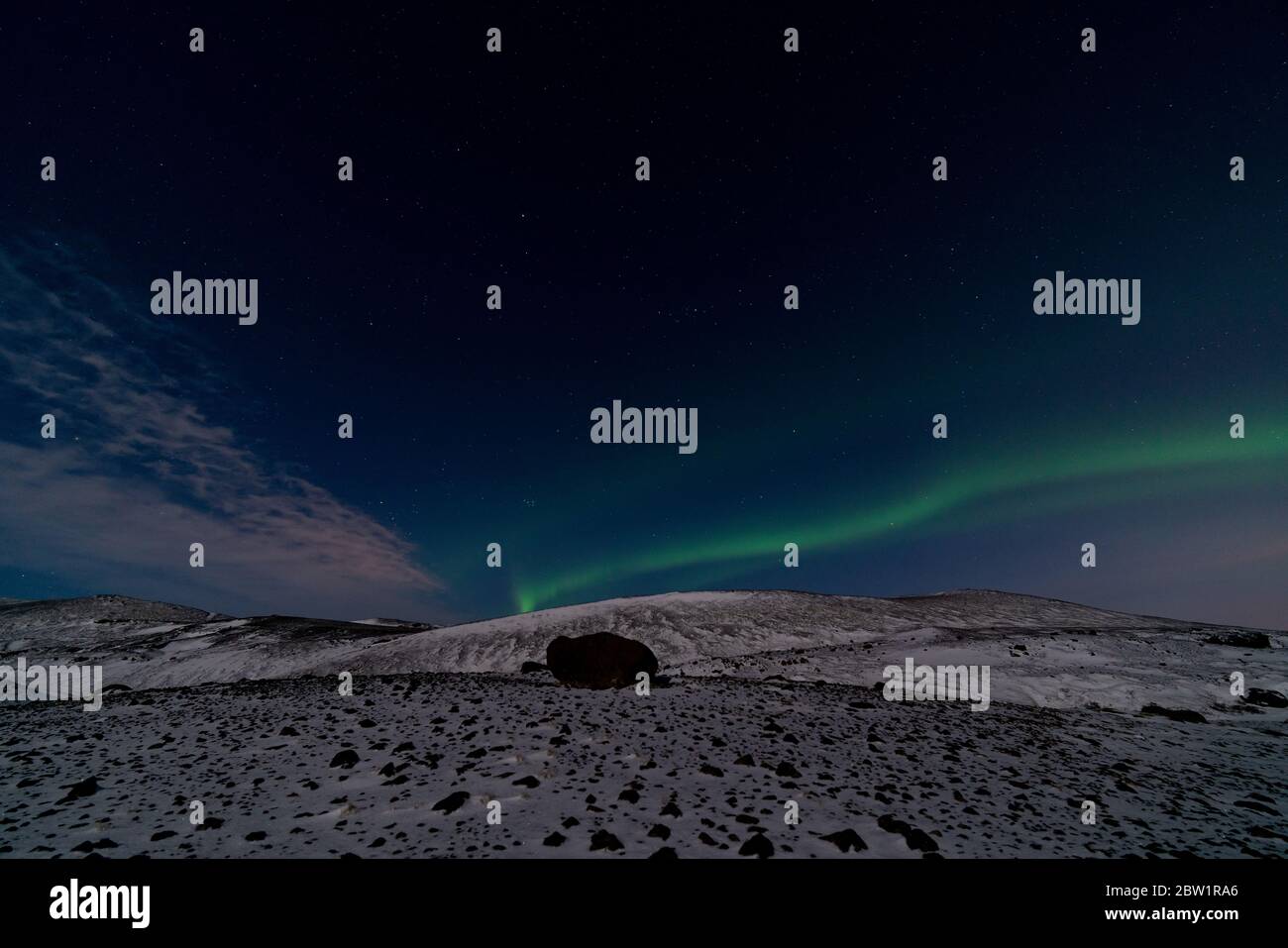 Nordlichter in Island am dunkelblauen Himmel mit einem Hauch von Wolken über einer schneebedeckten kargen Berglandschaft mit einem großen Felsen im Vordergrund. Stockfoto