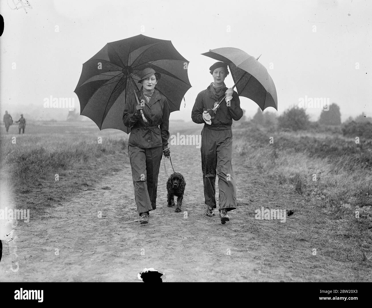 Lady Waleran (links) und Mrs locker Lampson tragen wasserdichte Hosen und tragen Regenschirme auf dem durchnässten Golfplatz in Walton Heath, wo sie Henry Cotton (Großbritannien) und Densmore Shute (USA) beim WM-Spiel zuschauen. 12 Juli 1937 Stockfoto