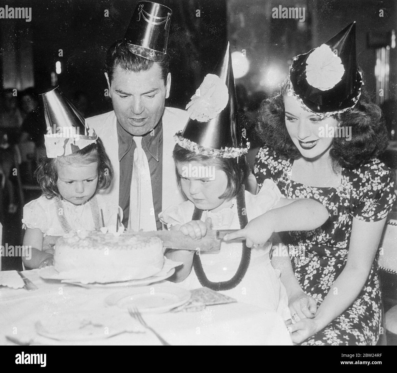 Dempsey 'refert' Kuchen Schneidezeremonie auf Tochter Geburtstagsfeier. Jack Dempsey, in Papier, und seine Frau, die ehemalige Hannah Williams, überwachen das Schneiden der Kuchen auf der Party gegeben, um den zweiten Geburtstag ihrer Tochter, Joan (rechts) zu feiern. Barbara, ihre andere Tochter, vier Jahre alt, führt die Zeremonie im Restaurant Dempsey's Broadway (New York) auf. September 1938 Stockfoto