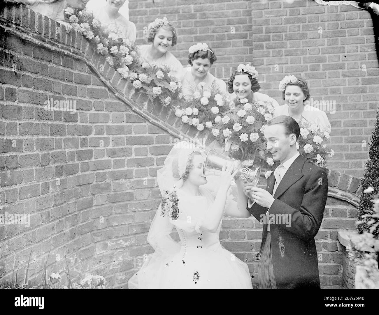 Besitzer des Hundderby Gewinner ist verheiratet in Brompton Oratory. Jack Walsh, der Besitzer von Lane keel, Gewinner des Greyhound Derby, war in Brompton Oratory mit Miss Ivy Sanders verheiratet. Alsh hat seiner Braut, Madelast April, versprochen, dass er ihr den Greyhound Derby Cup als Hochzeitsgeschenk überreichen würde. Foto zeigt, die Braut trinkt einen Toast aus dem Greyhound Derby Cup, gehalten vom Bräutigam, und beobachtet von den Brautjungfern bei der Hochzeitsfeier. Juli 1938 Stockfoto