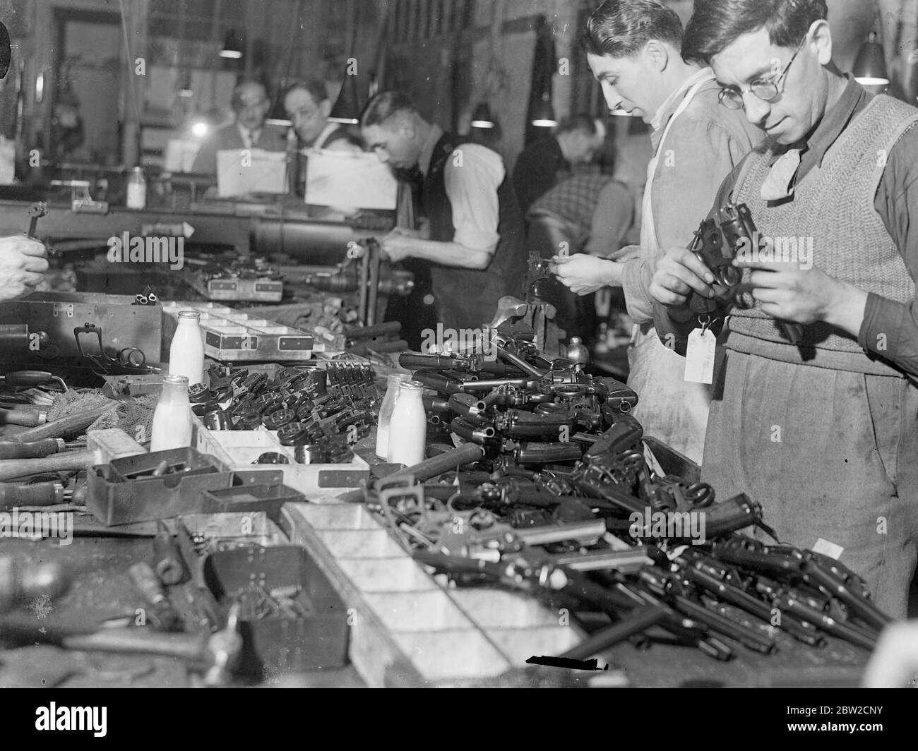 Zwischen drei und viertausend Flaschen Milch werden täglich von den Arbeitern in einer Fabrik konsumiert, die Tausende von Kleinwaffen wie Maschinengewehre und Revolver mit Massenproduktionsmethoden für das Versorgungsministerium produziert. Die Milch, die zu einem Penny pro Flasche verkauft wird, wurde für die Aufrechterhaltung der konstanten Leistung und die Senkung der Krankheitsrate unter den Arbeitern als unschätzbar befunden. Flaschen Milch auf der Bank im Revolverladen, wo Tausende von Kleinwaffen mit hoher Geschwindigkeit für die Verwendung der schnell wachsenden Kraft Großbritanniens ausgedreht werden. 15. November 1939 Stockfoto