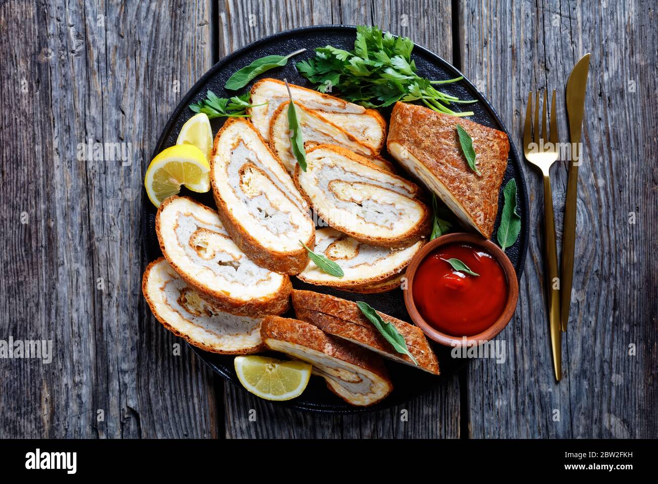 Gebackenes Omelett mit Hühnerfleisch auf einer schwarzen Platte serviert mit Zitrone, Tomatensauce und Petersilie auf grauem Holztisch mit goldener Gabel und kn Stockfoto