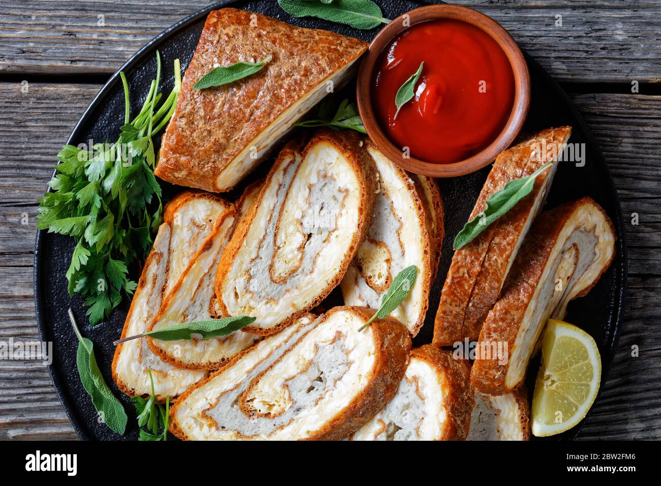 Nahaufnahme von Omelette Käse Roll mit Hackfleisch Füllung auf einem schwarzen Teller mit Zitrone, Tomatensauce und Petersilie auf grau rustikalen Holztisch, horizontal Stockfoto