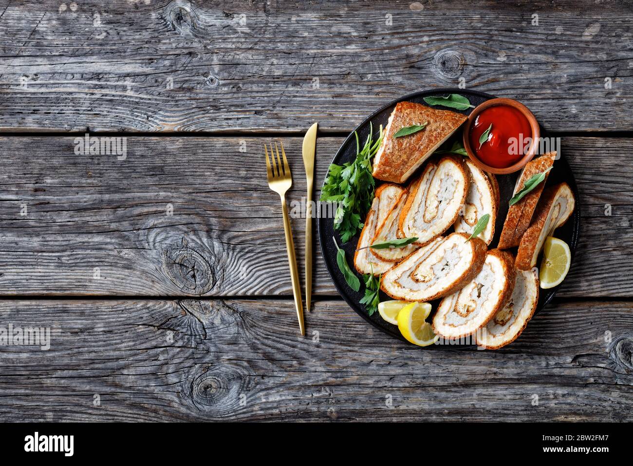 Omelette Käse Rolle mit Hackfleisch Füllung auf einem schwarzen Teller mit Zitrone, Tomatensauce und Petersilie auf grauen Holztisch mit goldenen Gabel und Messer, hor Stockfoto