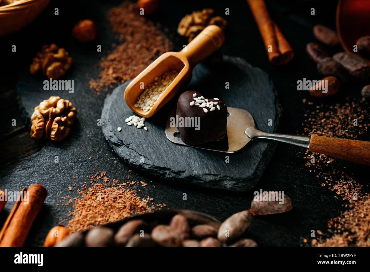 Schokolade Süßigkeiten auf Dessert Spatel in der Mitte von Kakaobohnen, Zimt, Nüsse, Kakaopulver. Nahaufnahme auf dunklem Stein Herz Form Hintergrund. SchokoladenDose Stockfoto