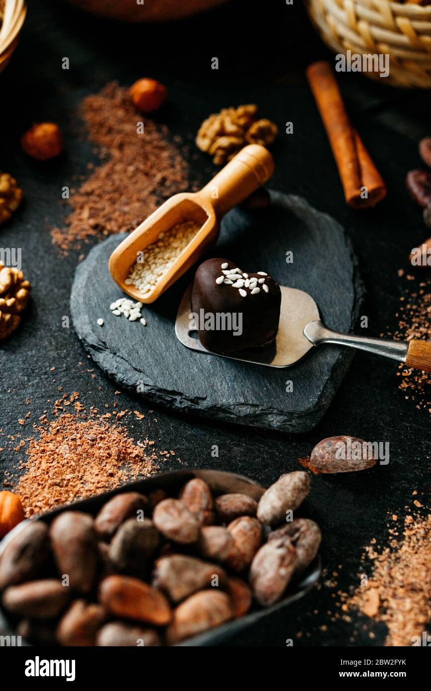 Schokolade Süßigkeiten auf Dessert Spatel in der Mitte von Kakaobohnen, Zimt, Nüsse, Kakaopulver. Nahaufnahme auf dunklem Stein Herz Form Hintergrund. SchokoladenDose Stockfoto