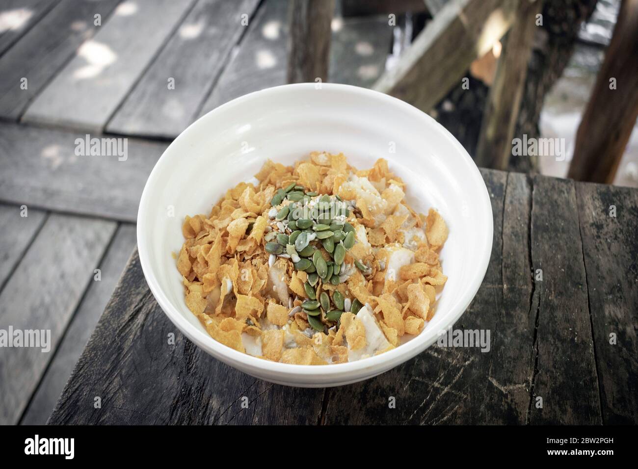 Gesundes Frühstück Obstschale mit Banane, Cornflakes, Kürbiskerne und Kokosmilch auf rustikalem Holztisch im Freien Stockfoto