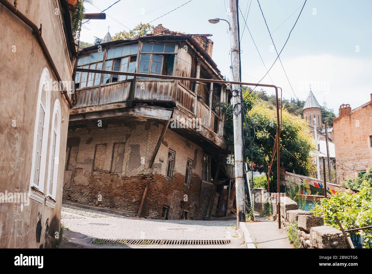 Ein schiefe, zerbröckelnde Holz- und Ziegelhaus in Tiflis, Georgien. Das Rohr, das im rechten Winkel über die Straße gebogen wird, transportiert Erdgas Stockfoto