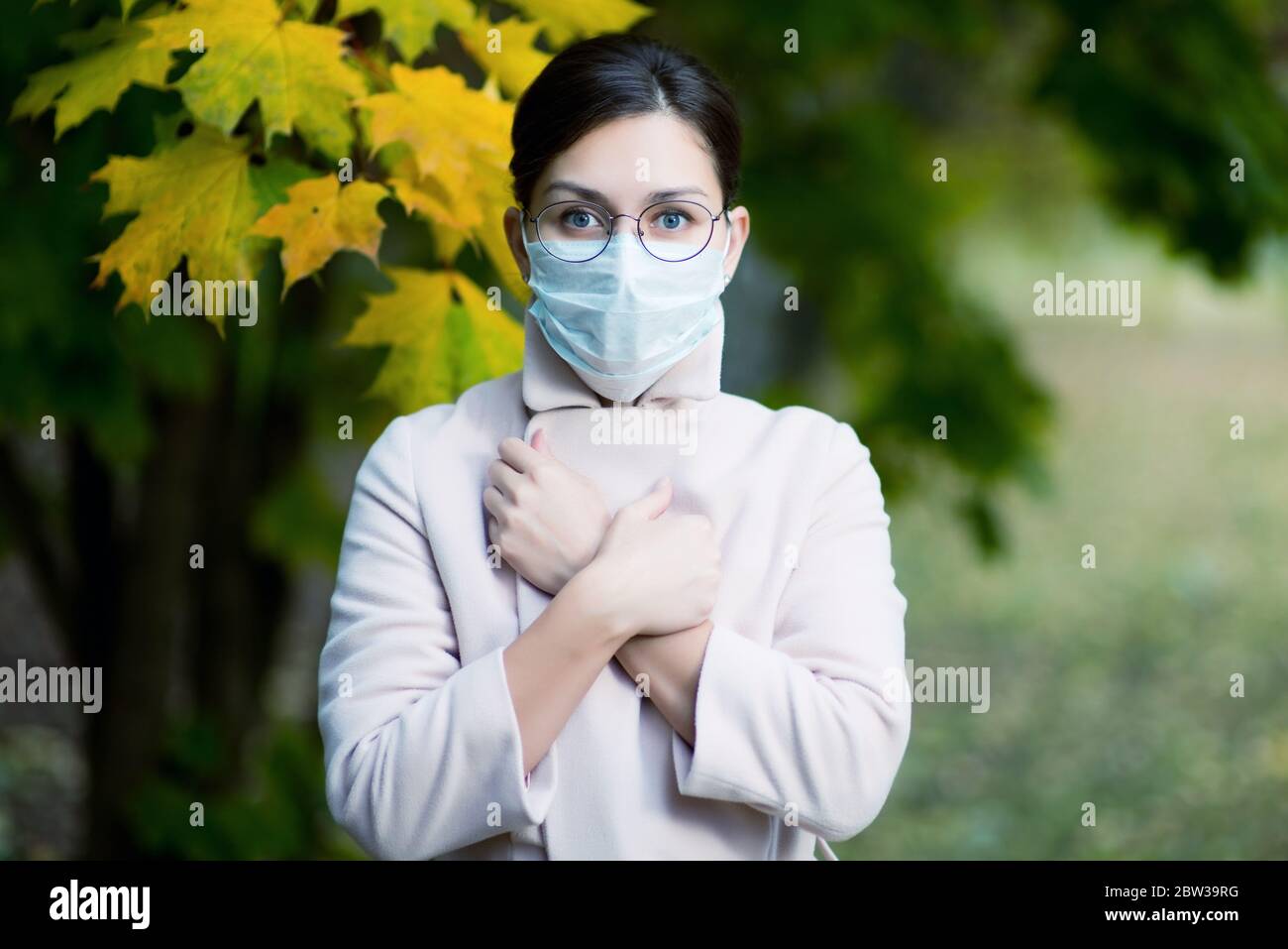 Eine junge Frau in Gesichtsmaske und Brille zum Sehen, umhüllt sich in einen Mantel und blickt mit weit geöffneten Augen vor sich. Kühle Farbe Stockfoto