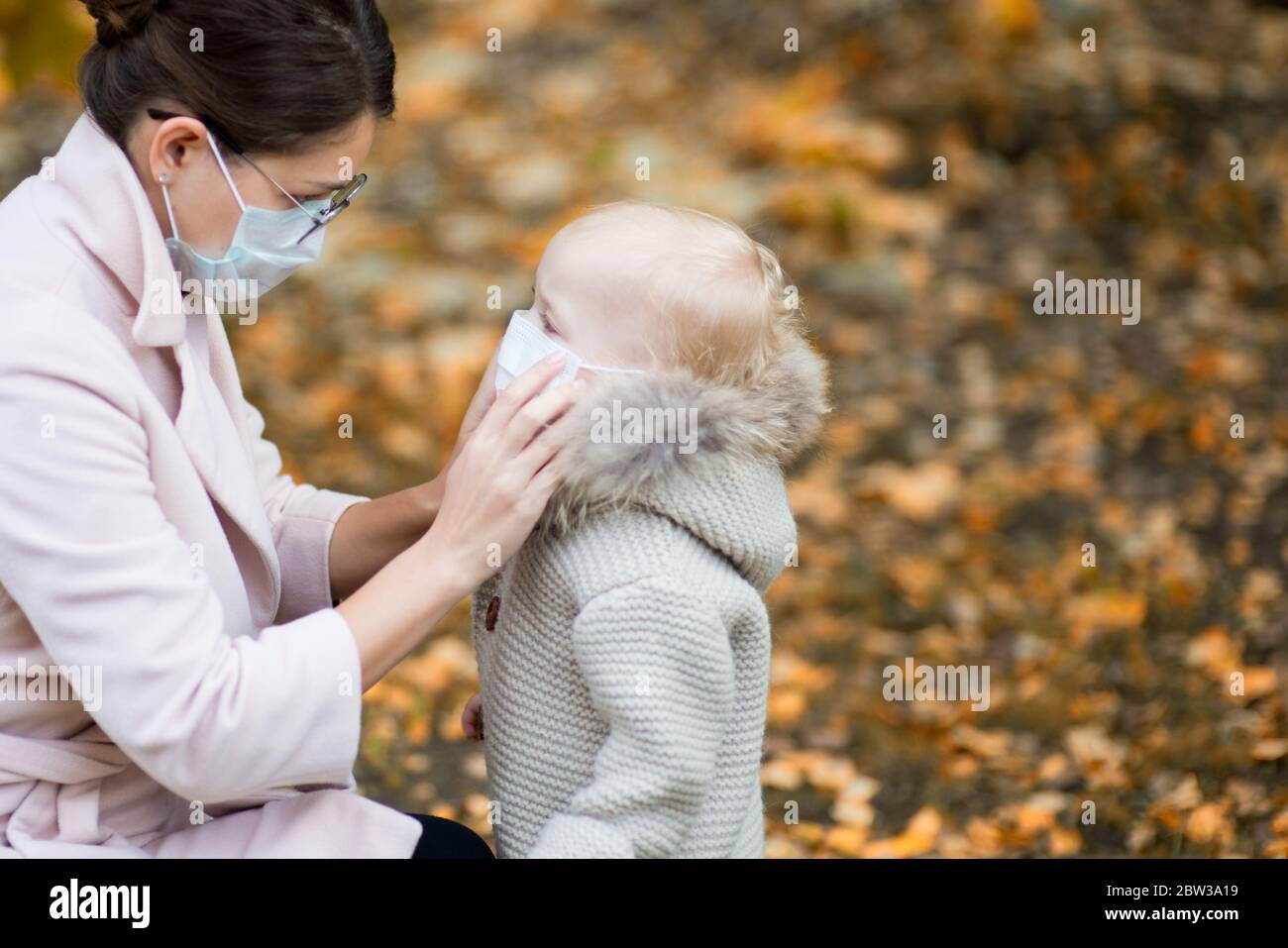 Mutter in medizinischer Gesichtsmaske und Brille passt die medizinische Maske vor dem Hintergrund des Herbsthofs ihrer kleinen Tochter an Stockfoto