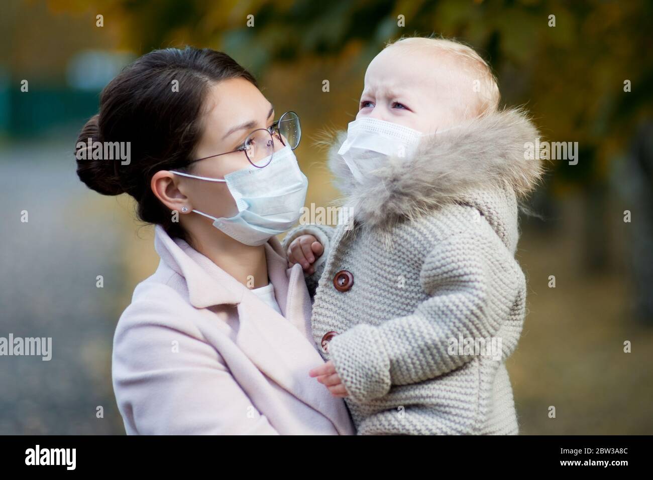 Mama in einer medizinischen Gesichtsmaske hält eine weinende Tochter in ihren Armen nah auf Stockfoto