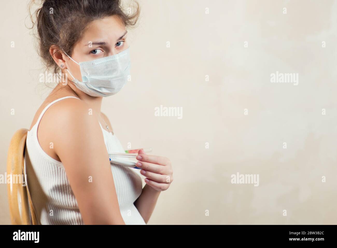 Ein kränklich aussehendes Mädchen in einer schützenden medizinischen Maske schaut traurig auf die Kamera, sitzt auf einem Stuhl mit mehreren Thermometern unter ihrer Achselhöhle Stockfoto