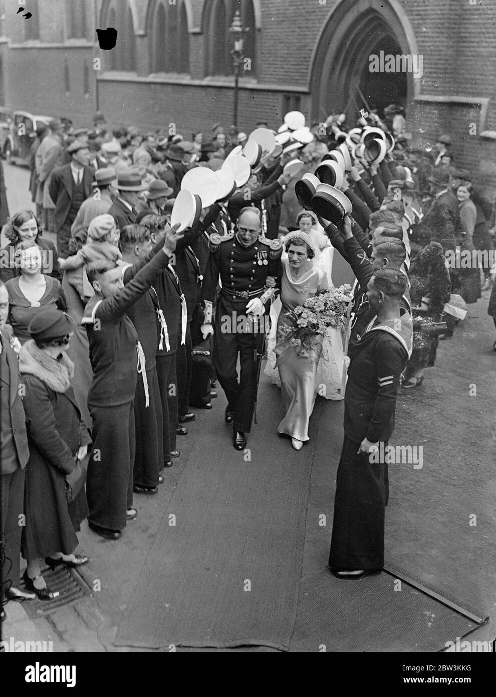 Marinewache der Ehre über die Straße bei der Hochzeit in London. Offiziere und Männer der HMS Iron Duke bildeten eine Wache der Ehre, die sich über die Straße, wenn die Hochzeit fand in ST Mary ' s Kirche, Cadus Street von Leutnant Commander Sydney Horn R N, und Miss Rita Mollins, Verkehr wurde gehalten. Foto zeigt, die Braut und Bräutigam unter dem Torbogen der Matrosen Hüte vorbei. Mai 1936 Stockfoto