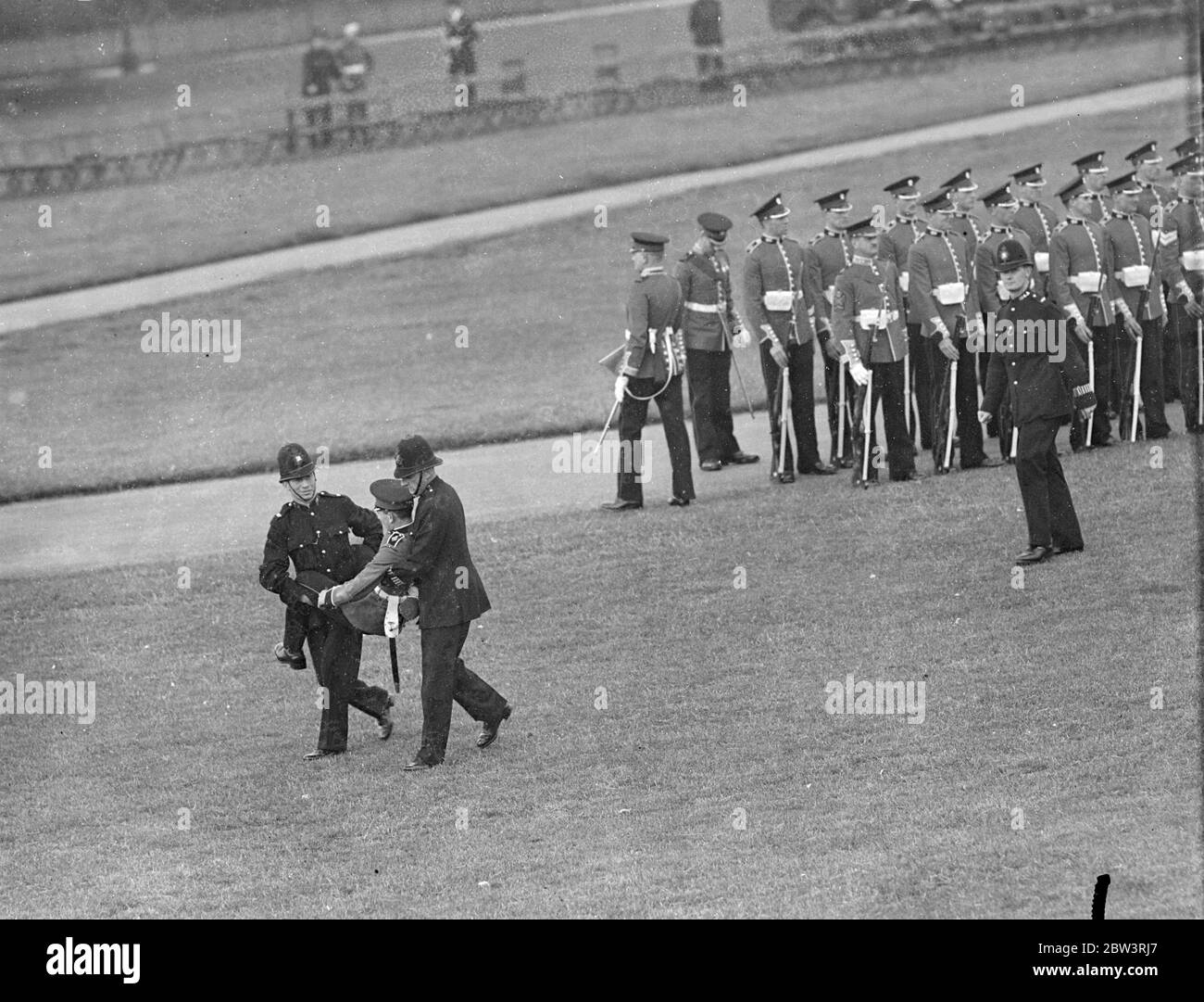 Guardsman ohnmächtig bei der Präsentation der Farben Probe . Ein Wachmann war im Hyde Park übertrieben, als am kommenden Donnerstag die Probe für die Präsentation der Neuen Farben für das Garde-Regiment vom König abgehalten wurde. Er wurde von Polizisten getragen. Foto zeigt, der Wachmann wird von zwei Polizisten mitgenommen. 14 Juli 1936 . Stockfoto