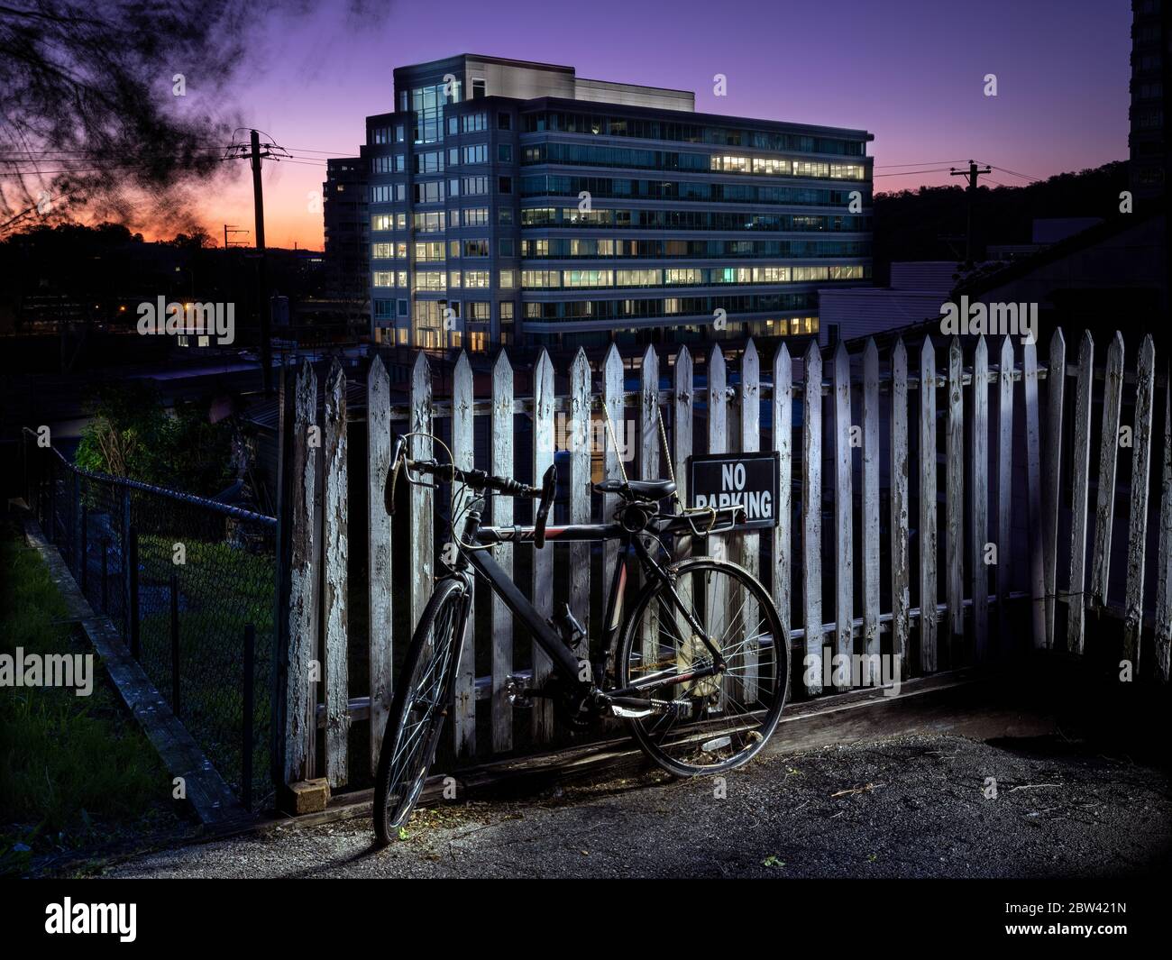 Altes Fahrrad & weißer Zaun in der Nacht mit modernem Bürogebäude im Hintergrund Stockfoto