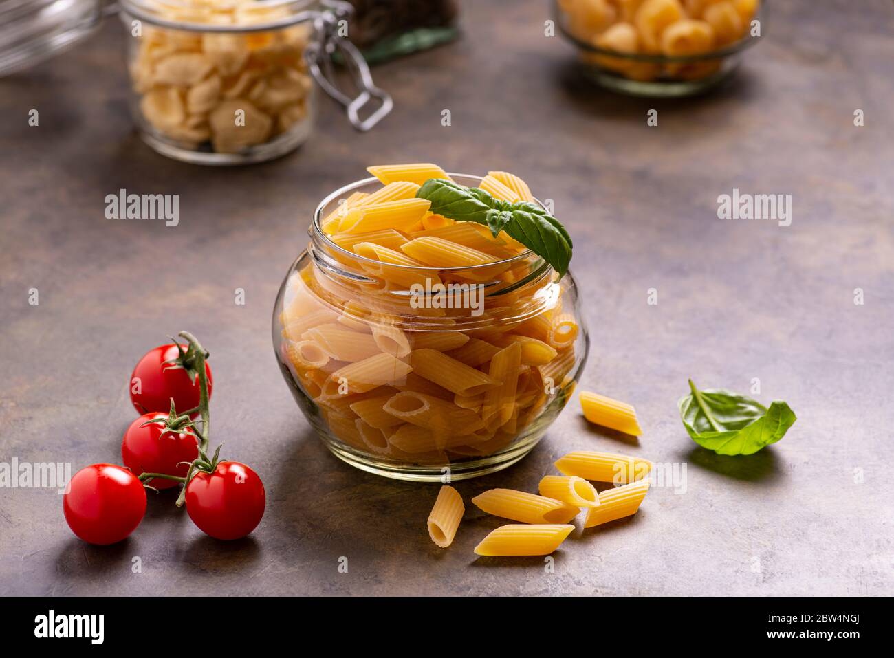 Im Vordergrund in einem transparenten Glas rohe italienische Pasta und frische Basilikumblätter. Im Hintergrund ein Bündel Kirschtomaten Stockfoto