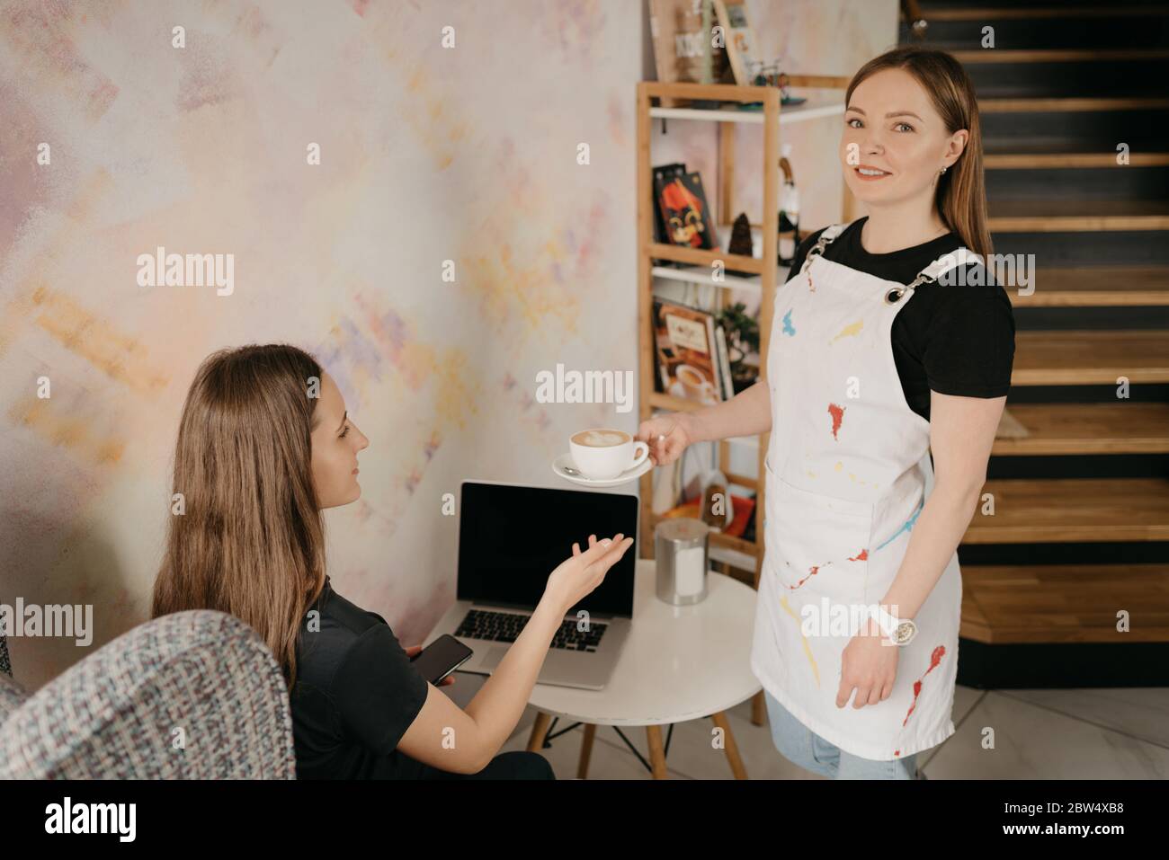 Eine Barista mit Gesichtserpiercing hält einem Mädchen in einem Café einen Latte vor. Eine junge Frau mit langen Haaren, die ferngesteuert an einem Laptop arbeitet, hält soci Stockfoto